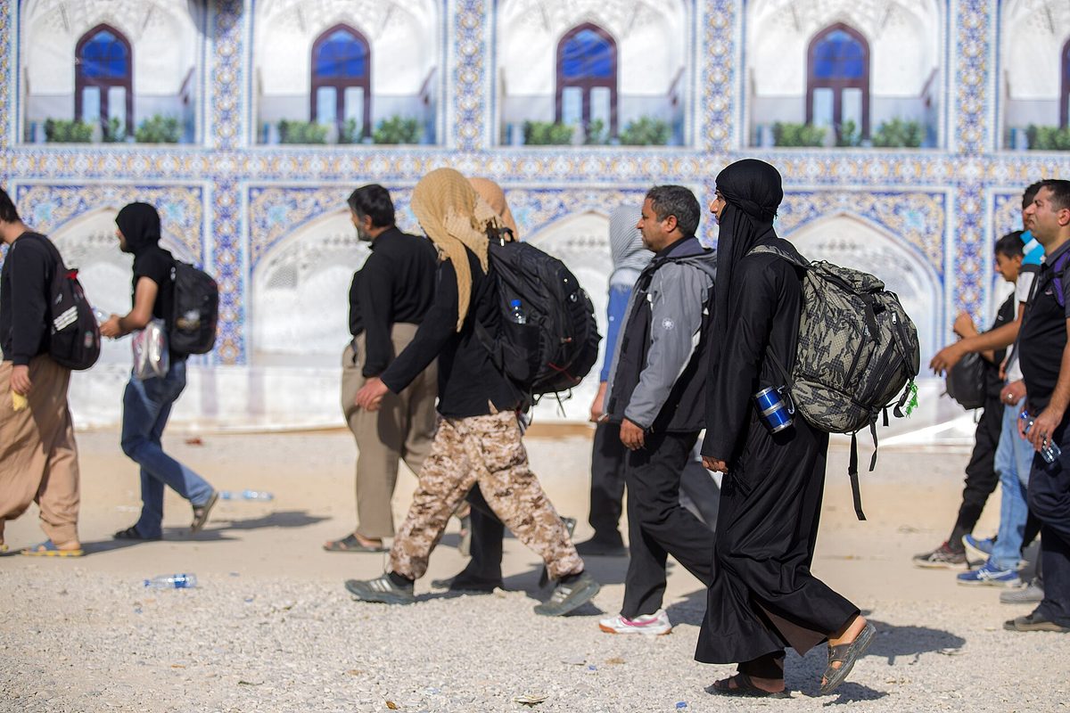 Arbaeen pilgrims with backpacks walking past shrine tilework on the route to Karbala