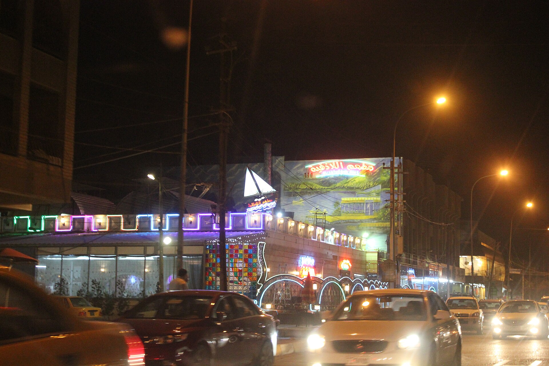 Brightly lit restaurant frontage with neon signage and traffic on Arasat al-Hindiya in Baghdad at night