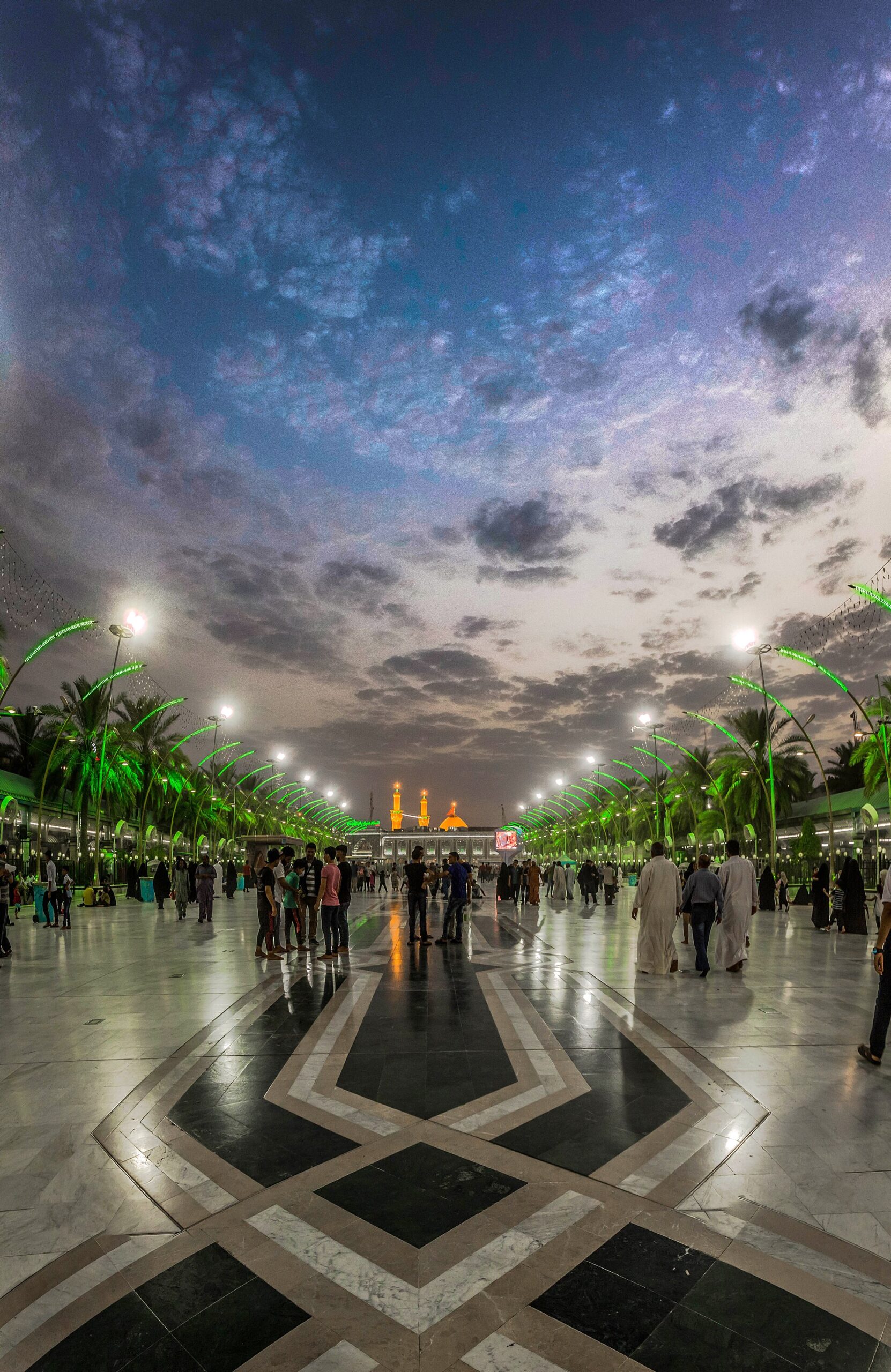 The paved Bayn al-Haramayn corridor between the Imam Hussein and al-Abbas shrines in Karbala, lit at dusk with palm trees along both sides