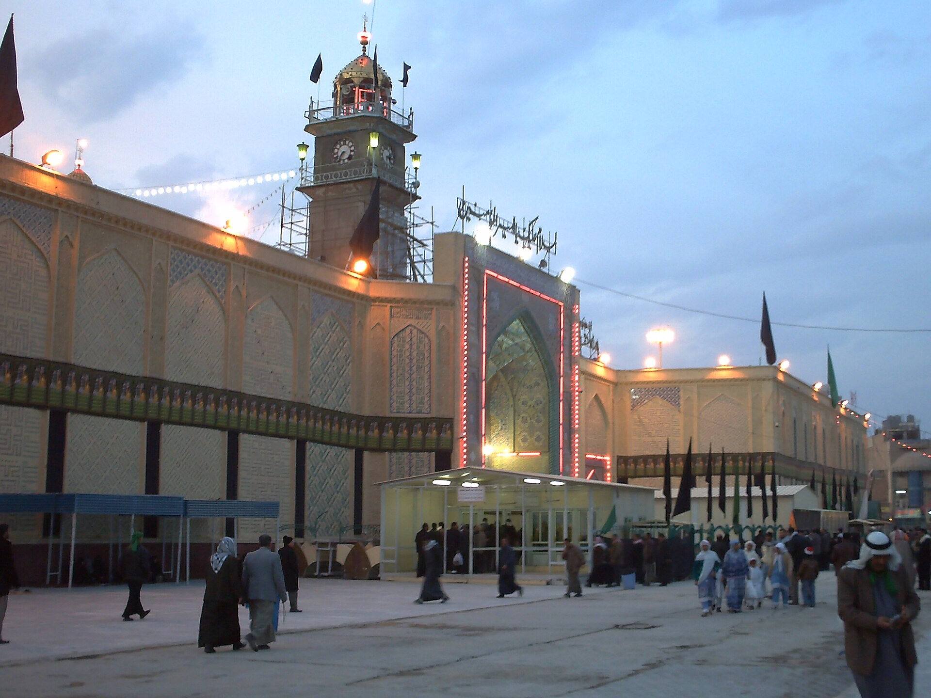 The outer plaza and clock tower of the Imam Ali shrine compound in Najaf at dusk with visitors crossing the open ground