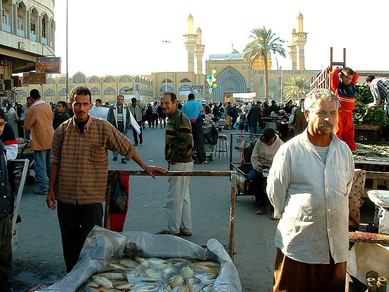Fish and vegetable stalls in a Kadhimiya street market with the shrine minarets rising behind