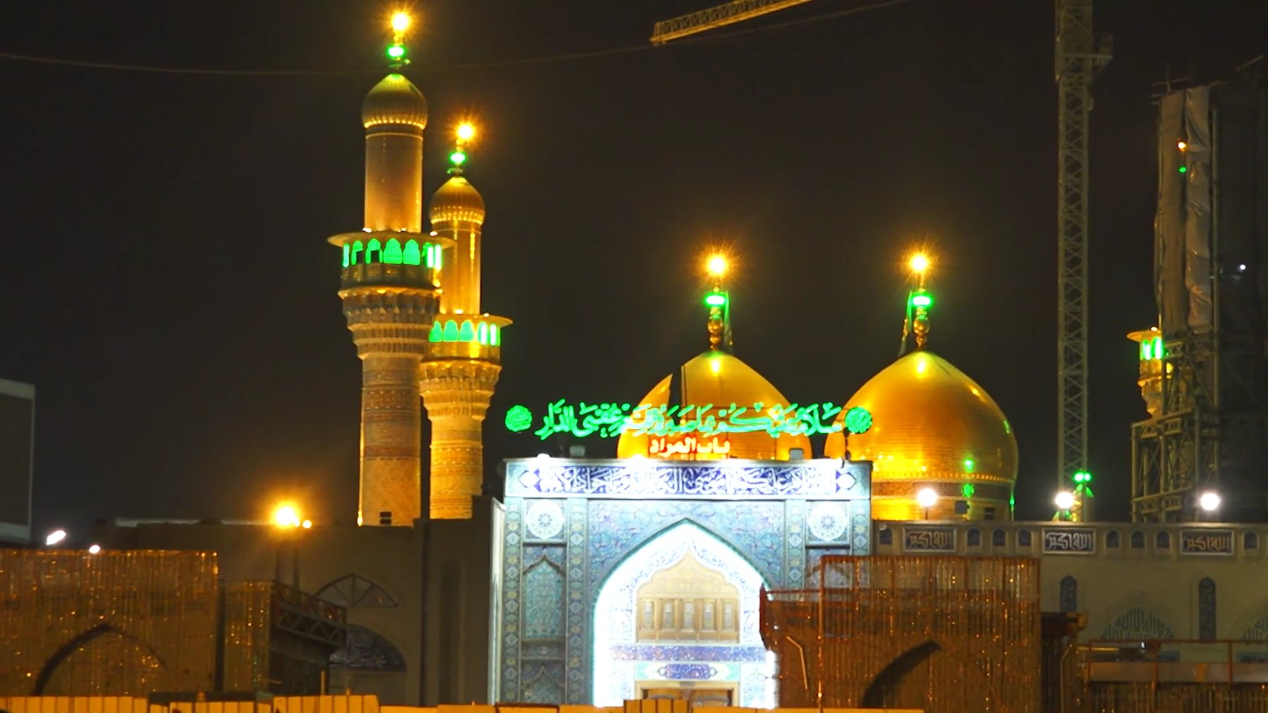 The twin gilded domes and gold minarets of the al-Kadhimiya shrine in Baghdad lit at night, with construction cranes from the expansion works