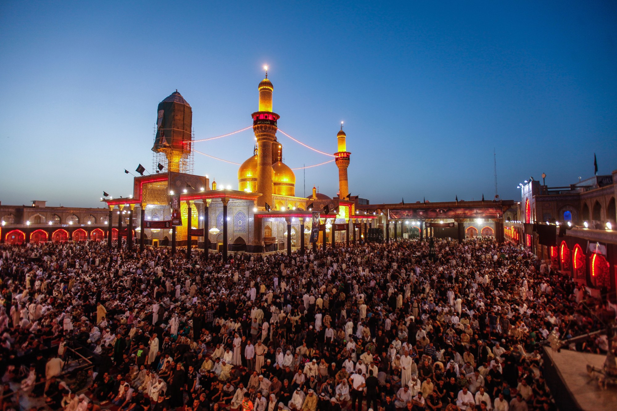 Crowds fill the courtyard of the Kadhimiya shrine at dusk beneath its twin gold domes and gold minarets