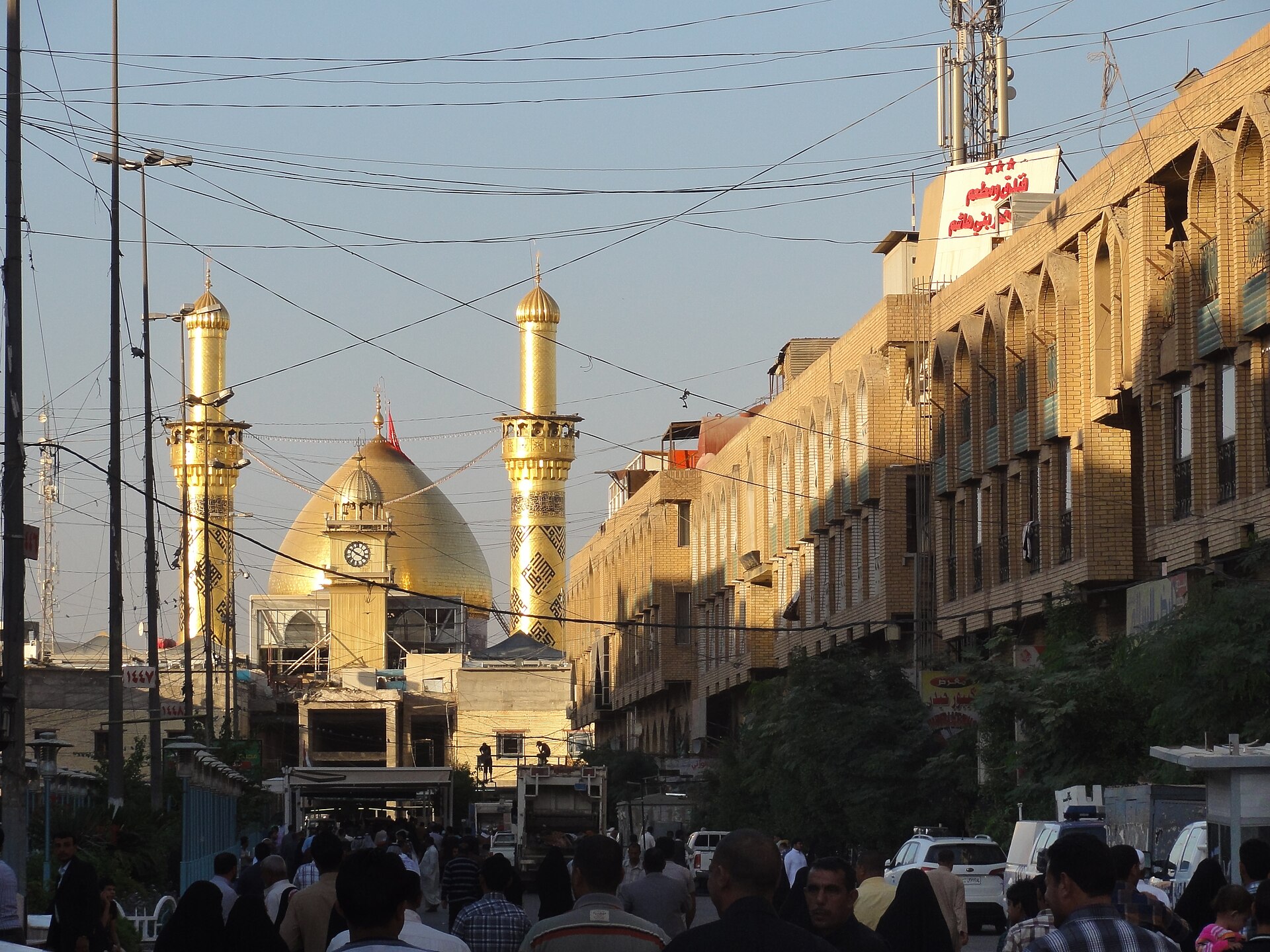 Street-level view in Karbala's haram district with the golden dome and minarets rising directly behind shopfronts and an arcaded commercial building