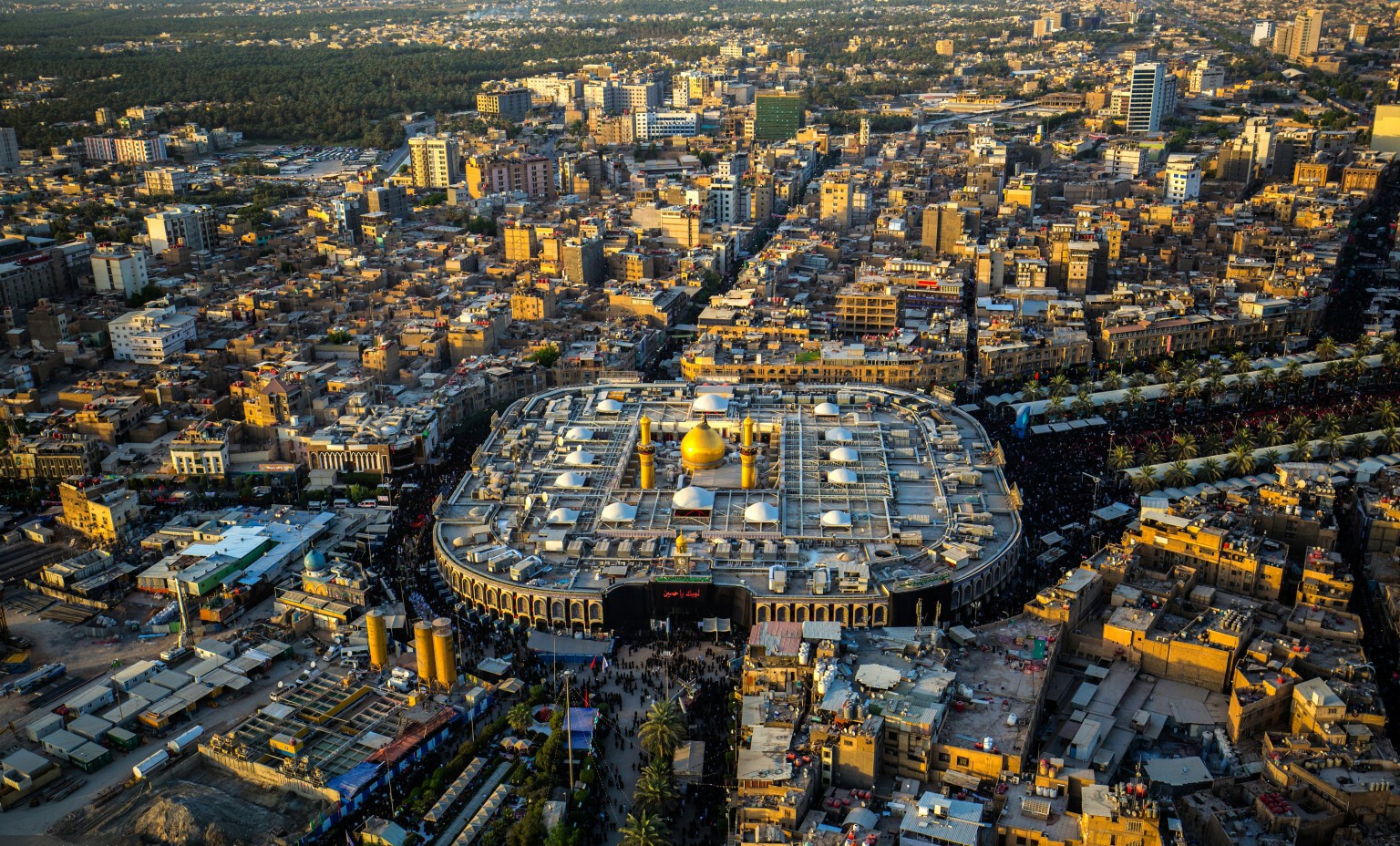 Close aerial view of a golden-domed shrine complex in Karbala ringed by tightly packed multi-storey buildings and market streets