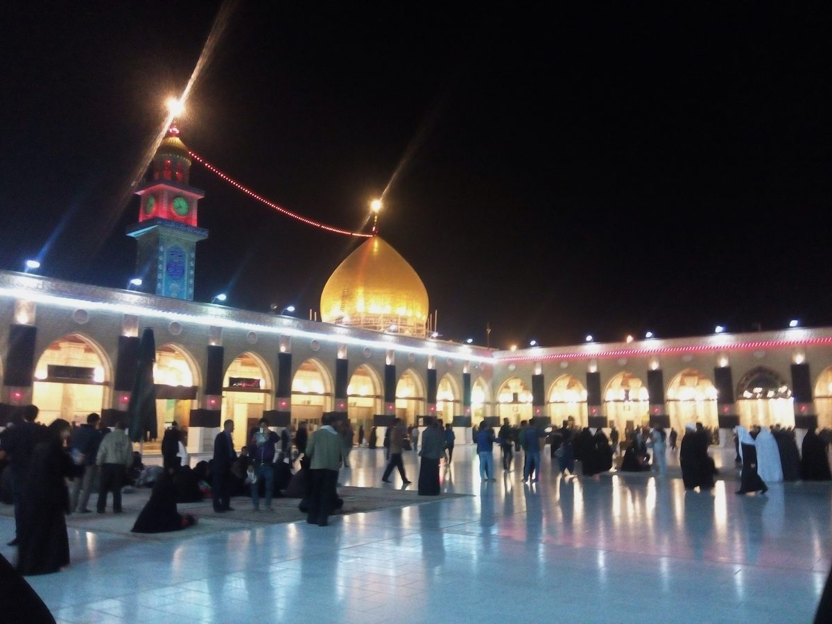 The Great Mosque of Kufa lit up at night with pilgrims crossing the courtyard