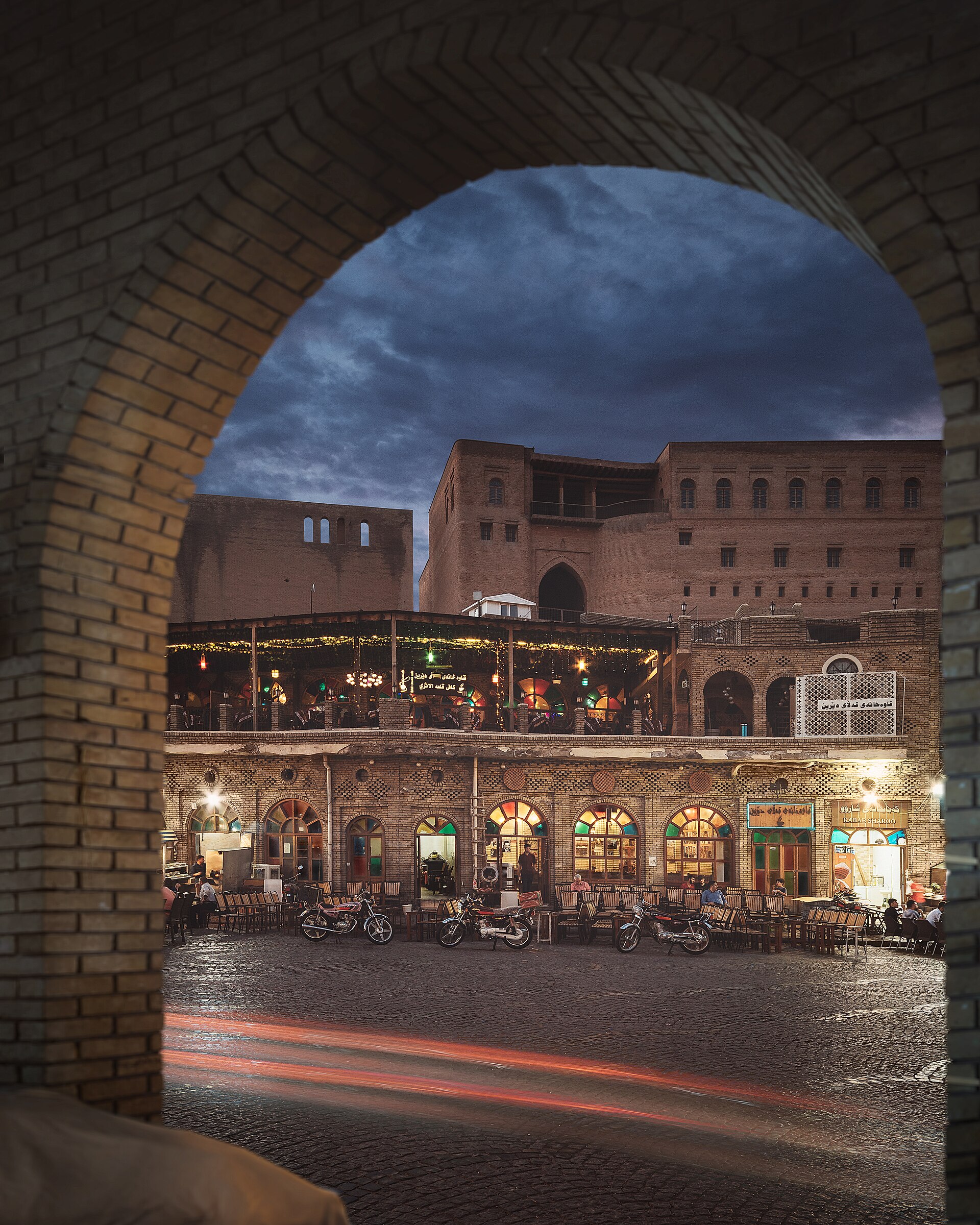 Teahouse tables and a lit upper terrace at the foot of the Erbil Citadel's southern wall at dusk