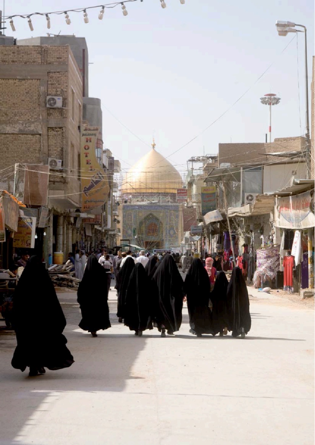 Pilgrims walking down Tusi Street in Najaf's old city towards the golden dome of the Imam Ali shrine, past shopfronts and a hotel sign