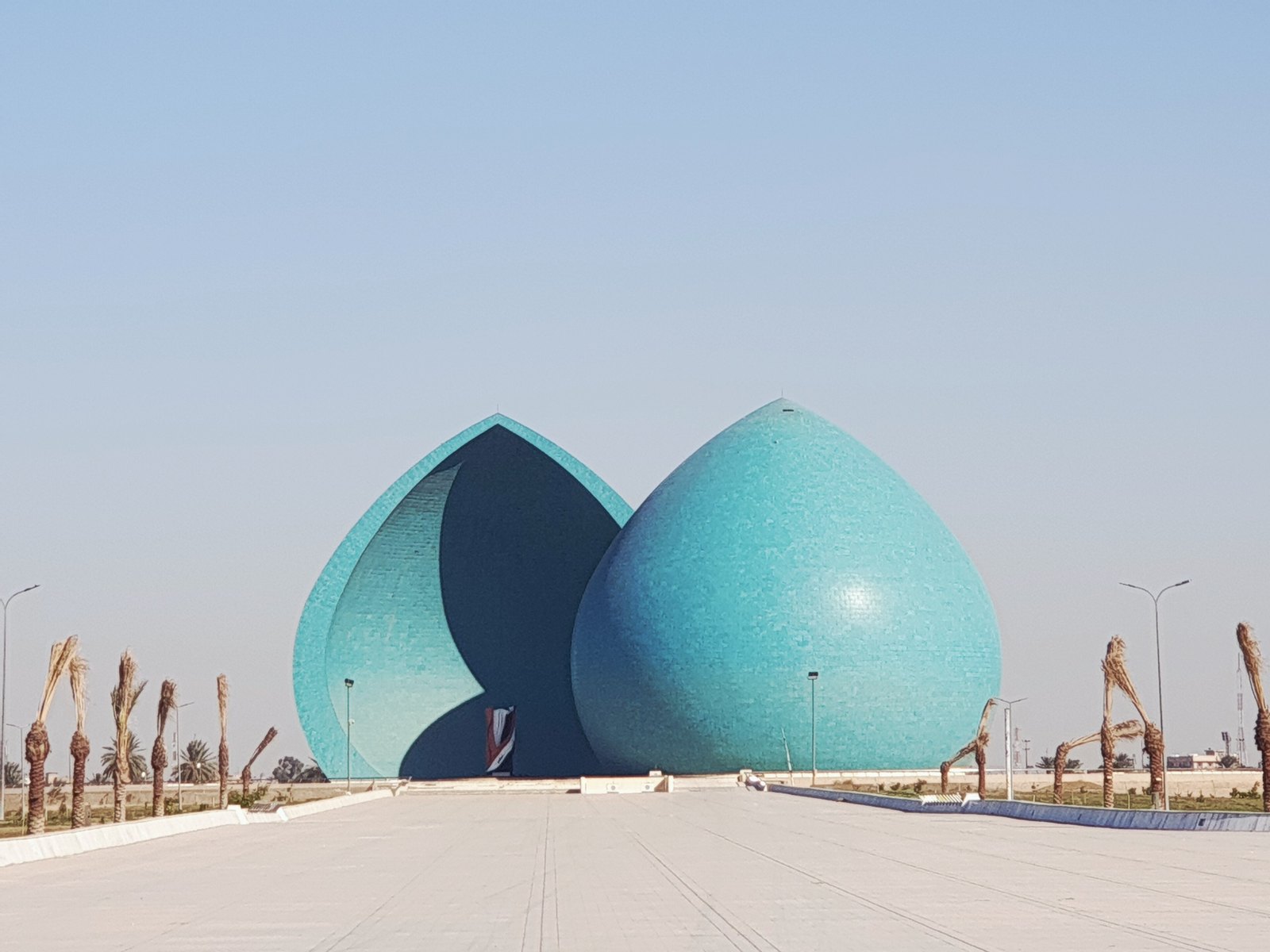 The two split turquoise half-domes of the al shaheed monument in Baghdad seen from the approach road, with the Iraqi flag sculpture between them
