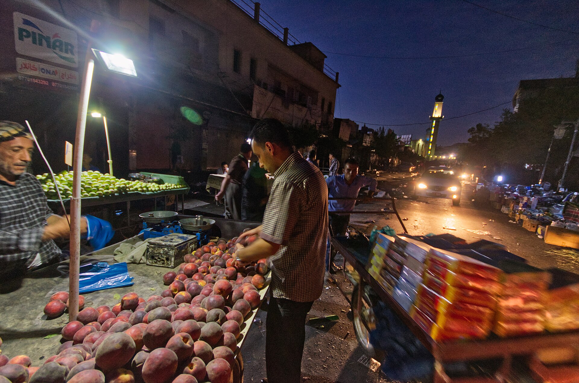 Fruit sellers working a street market in Sulaymaniyah after dark, with a minaret lit behind