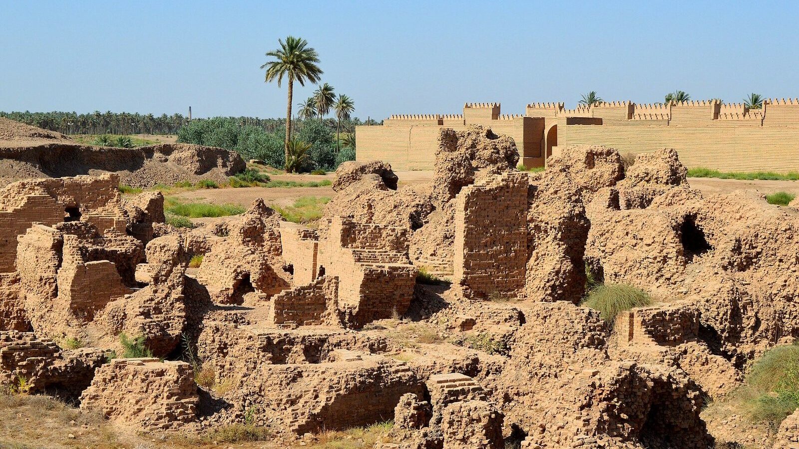 Mud-brick ruins of ancient Babylon in the foreground with reconstructed crenellated walls and palm trees behind