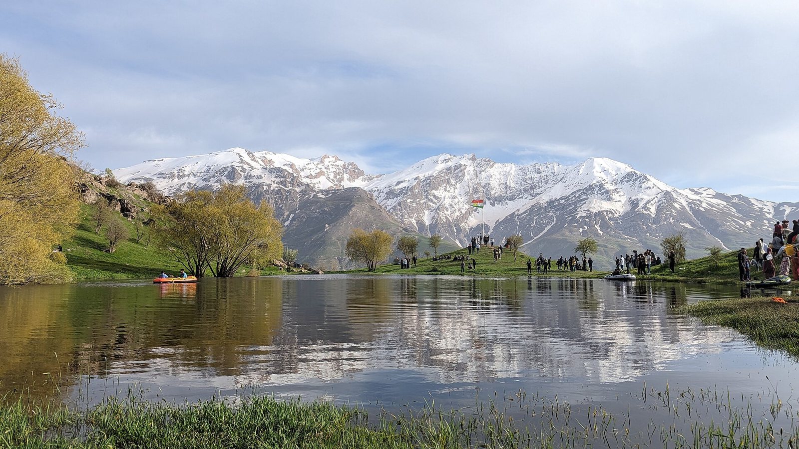 Visitors on the green shore of the Gomi Felaw lake near Choman with snow-covered mountains reflected in the water