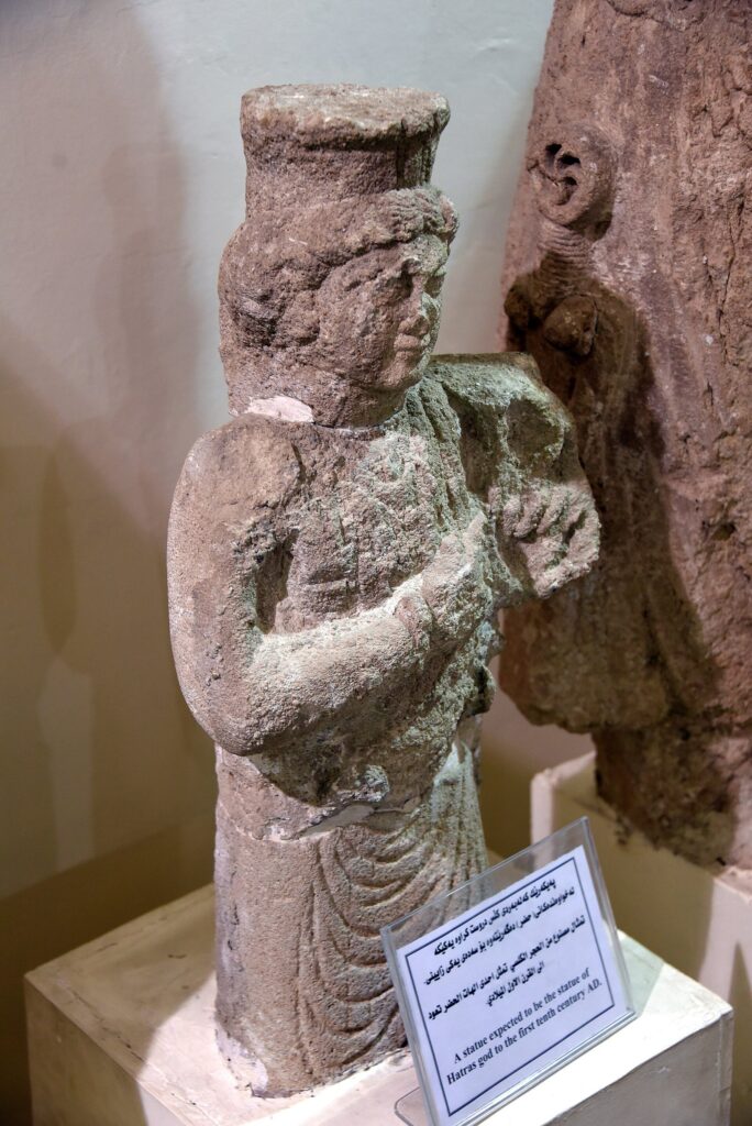 Weathered limestone statue of an unidentified goddess from the Great Temple at Hatra, in a museum case