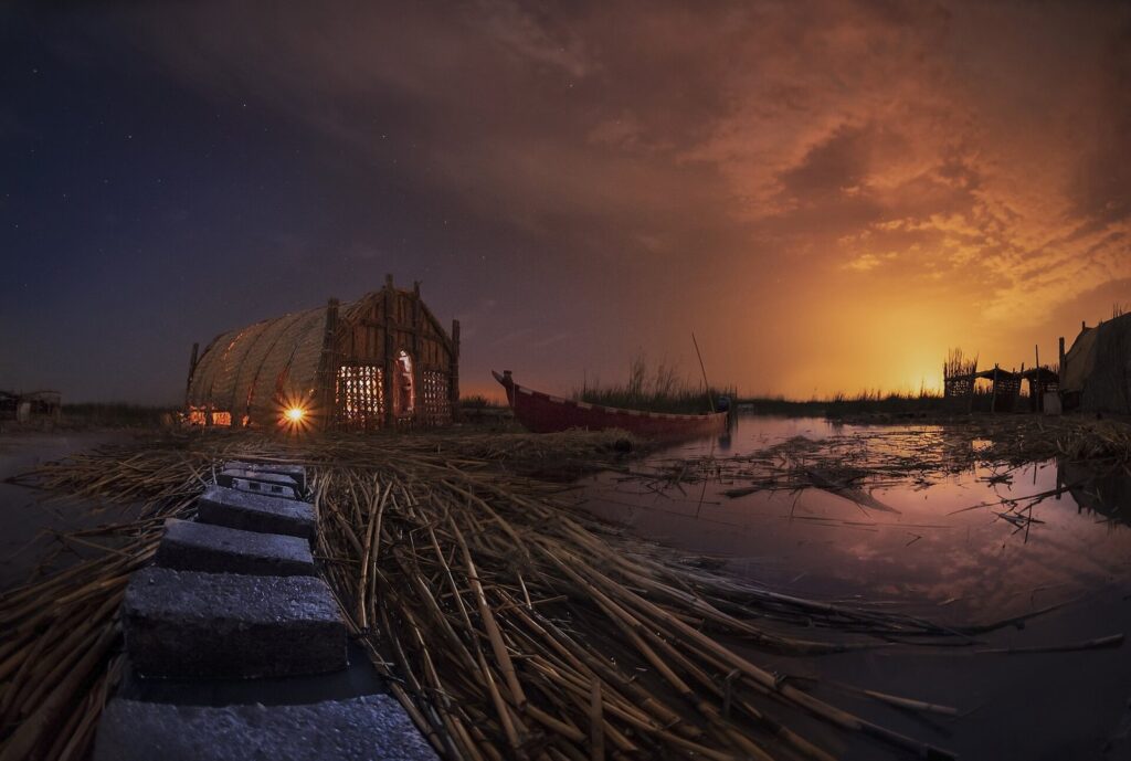 A reed mudhif guesthouse on the water of the Iraqi marshes at dusk, with a wooden walkway of reed bundles