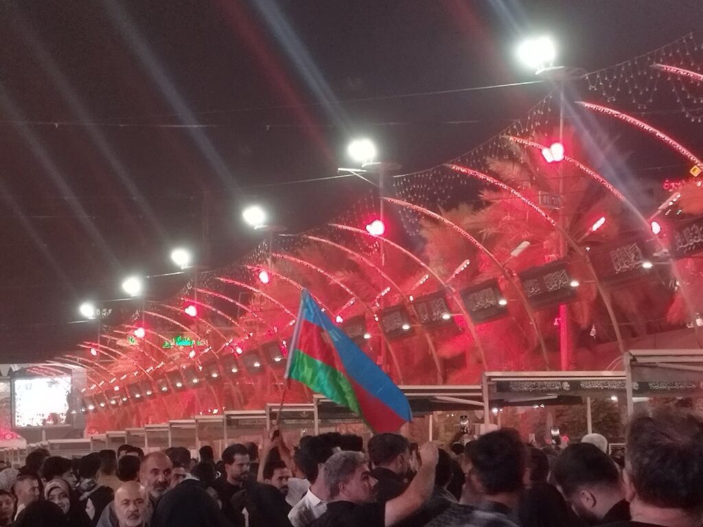 Pilgrims under the lit canopy of Bayn al-Haramayn in Karbala at night, one holding the flag of Azerbaijan