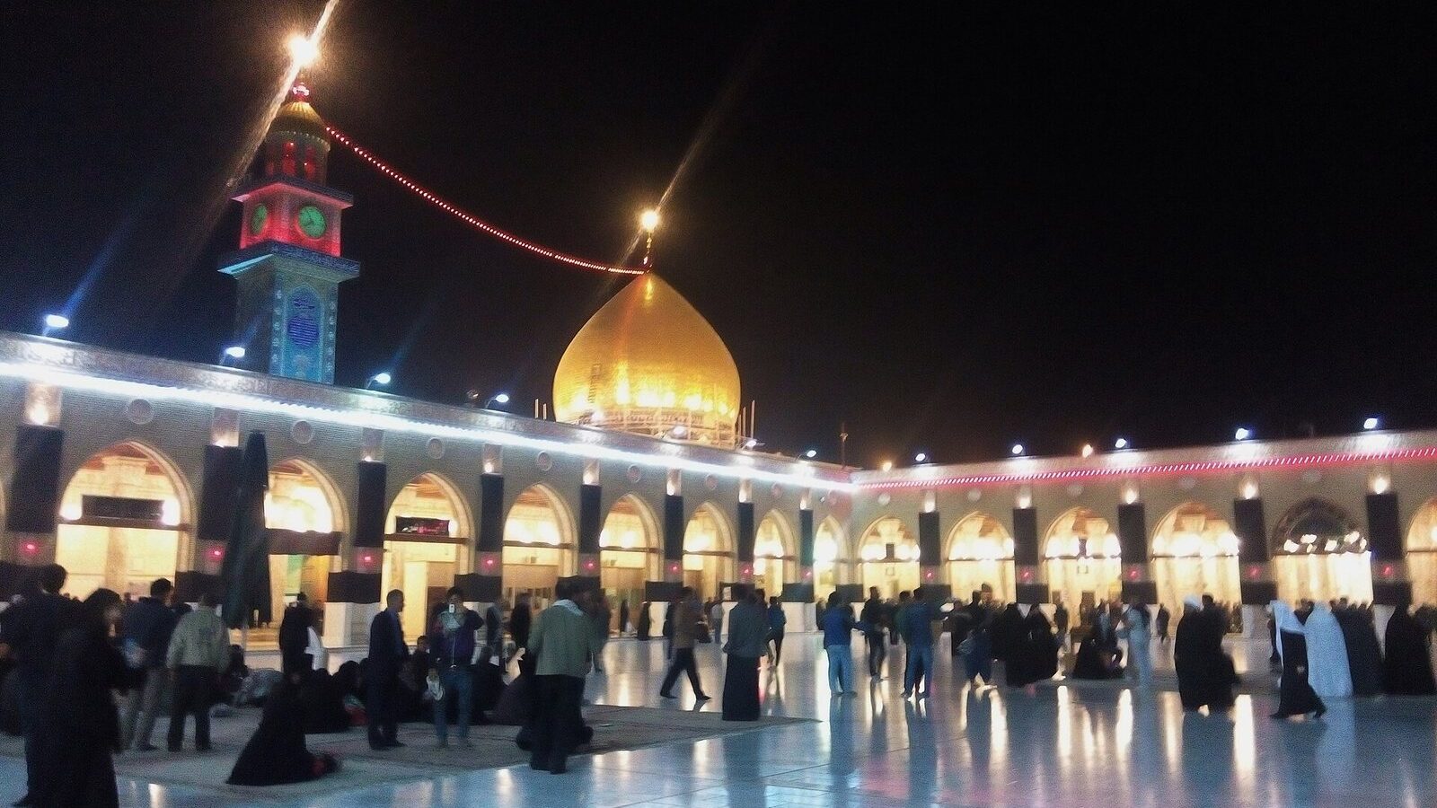 The lit courtyard and golden dome of the Great Mosque of Kufa at night with worshippers crossing the marble floor