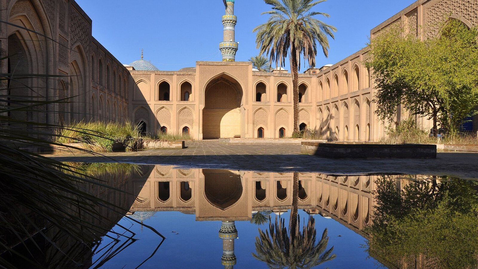 The arcaded courtyard of the Mustansiriya Madrasa in Baghdad reflected in a pool, with a minaret and palm tree