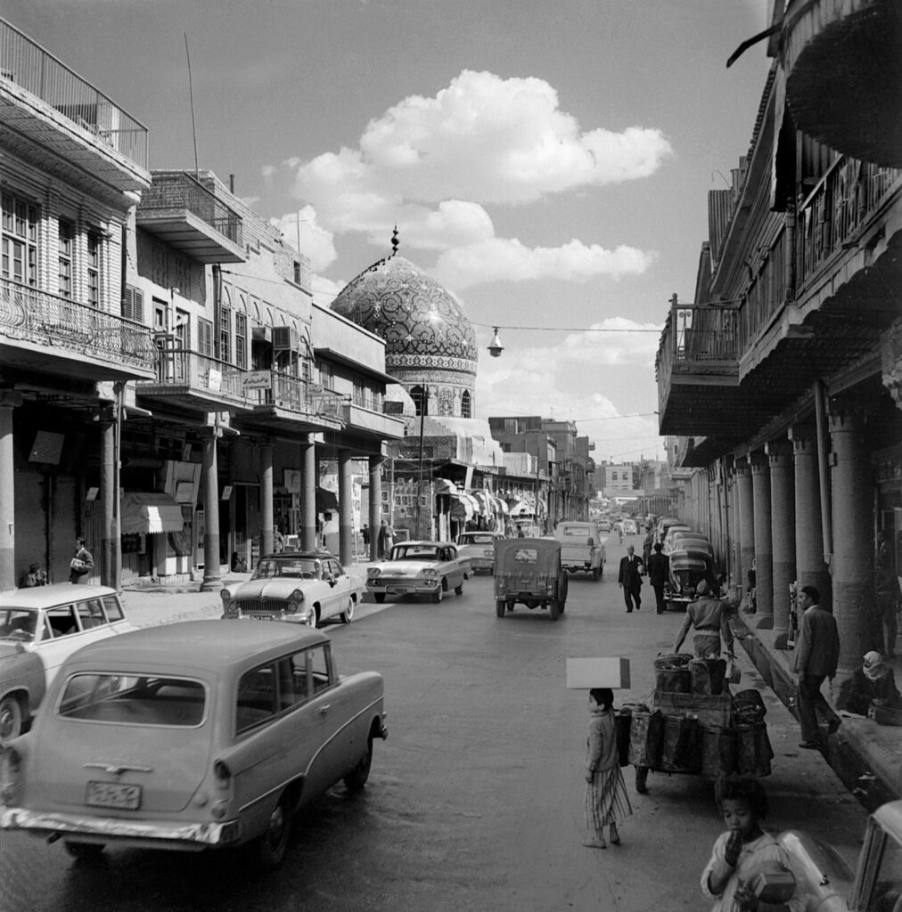 Rasheed Street in Baghdad in 1961 with colonnaded shopfronts, old cars and a tiled mosque dome in the distance